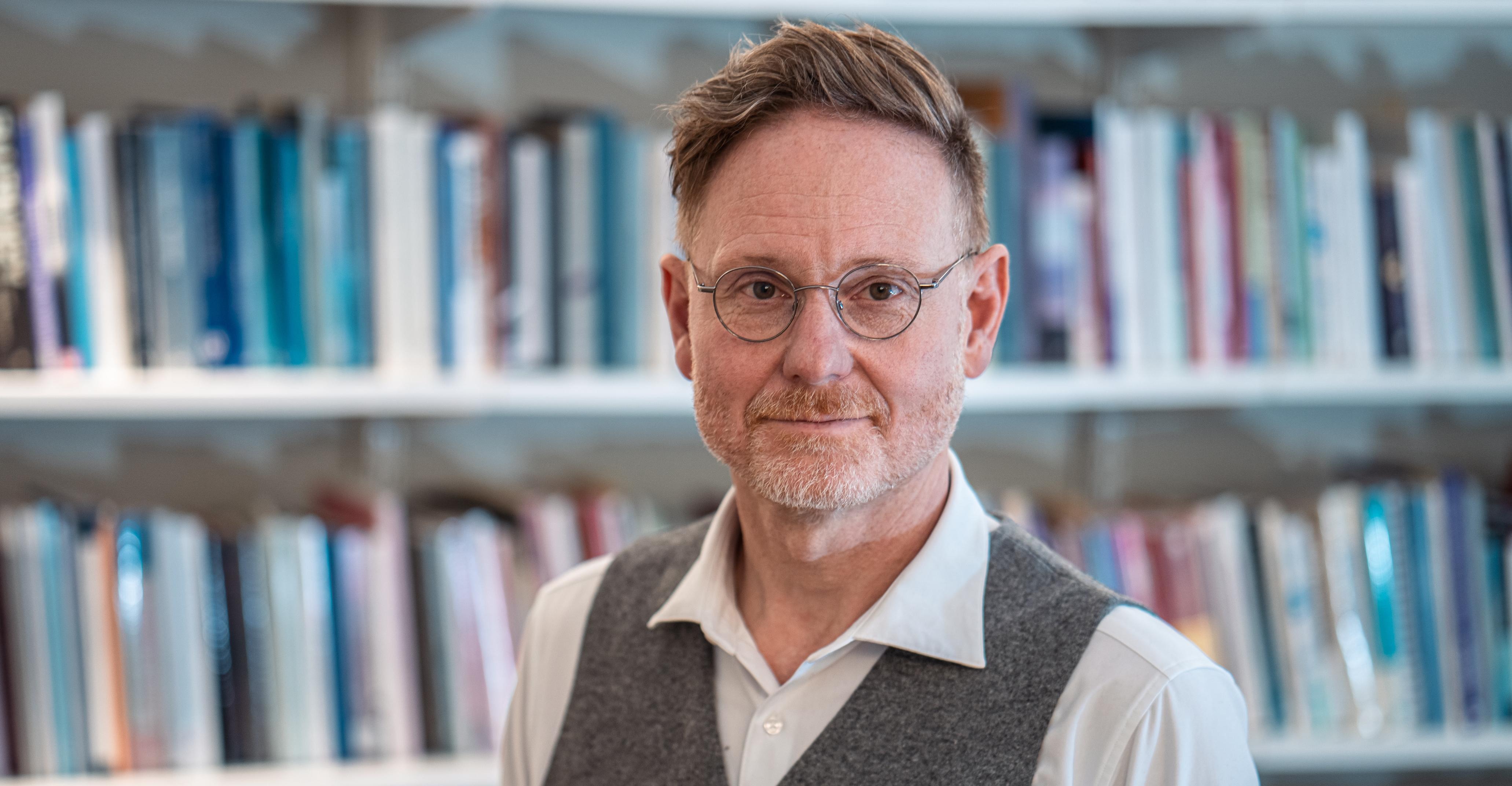 Magnus Karlsson har en vit skjorta, en grå väst och runda glasögon. Han tittar in i kameran och bakom honom syns många böcker i en vit bokhylla.