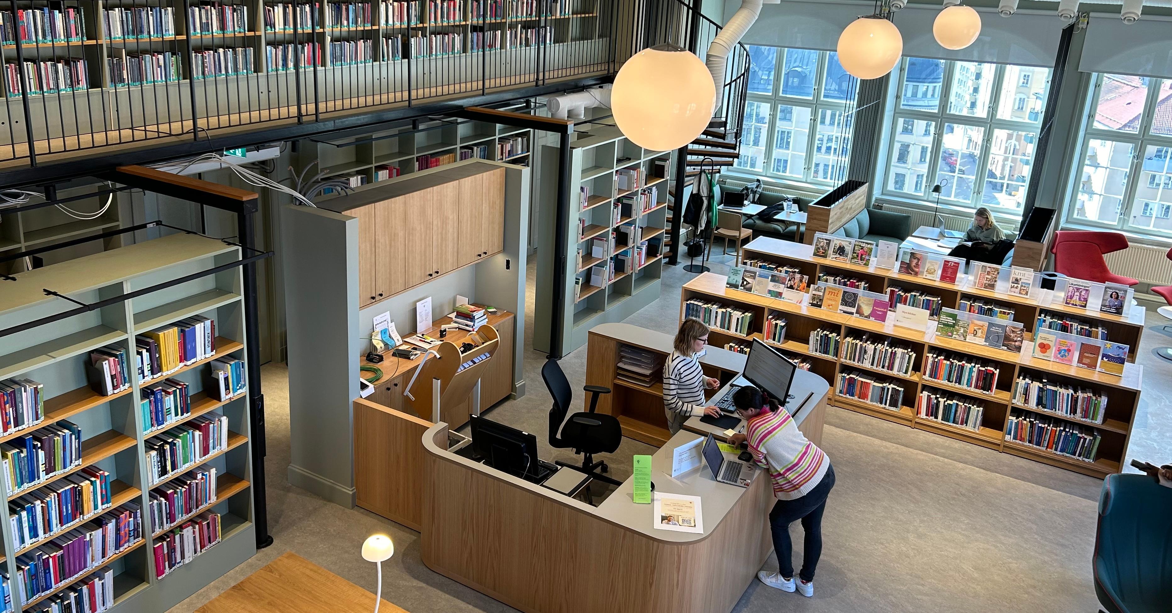 En överblicksbild i biblioteket där allting ses uppifrån: hyllor, bibliotekariedesken, studieplatser, fåtöljer och ett fåtal personer