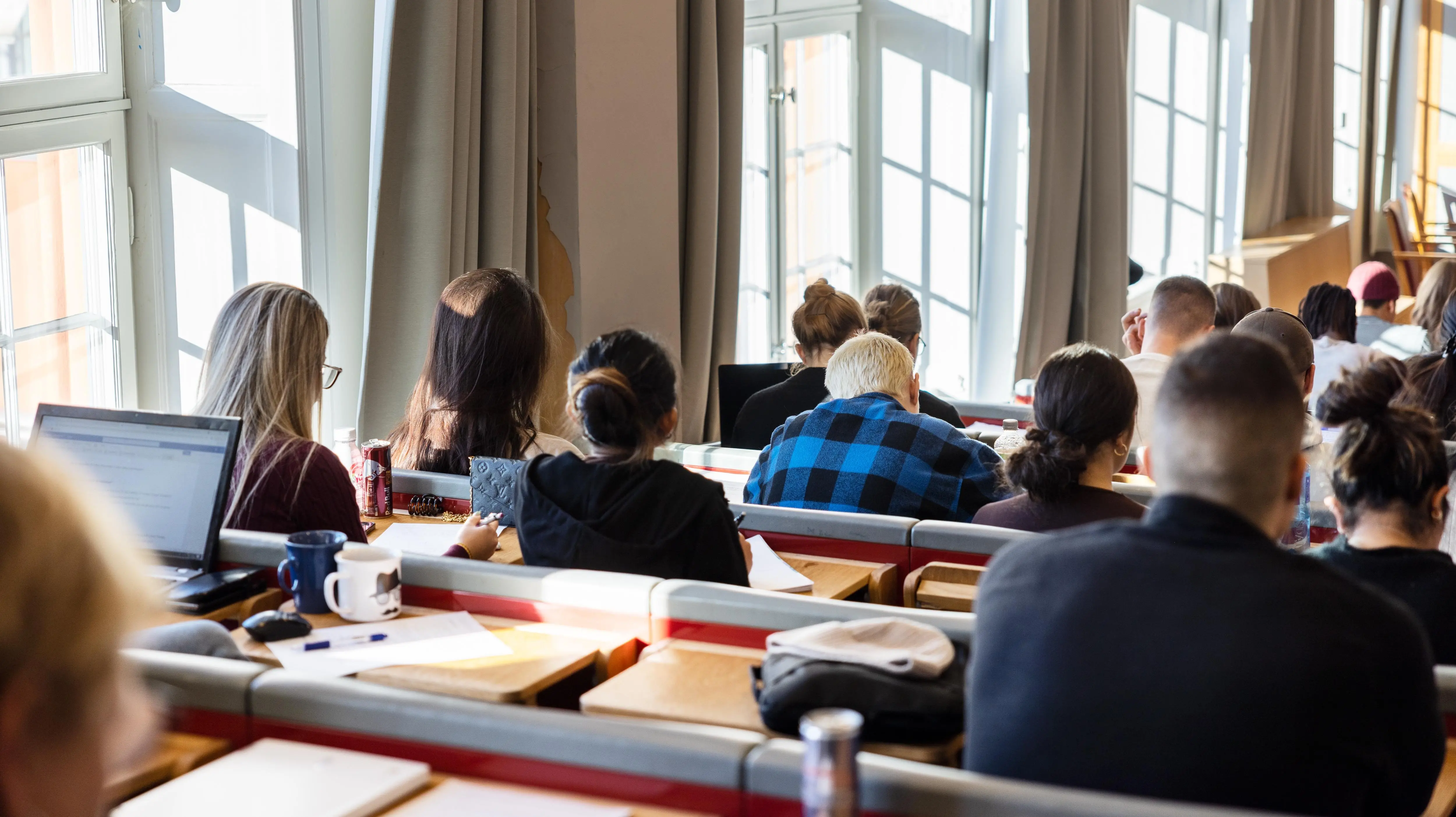 A large group of students are sitting with their backs against the camera in an auditorium while listening to a lecture. The room is bright and the sun is shining through the windows.