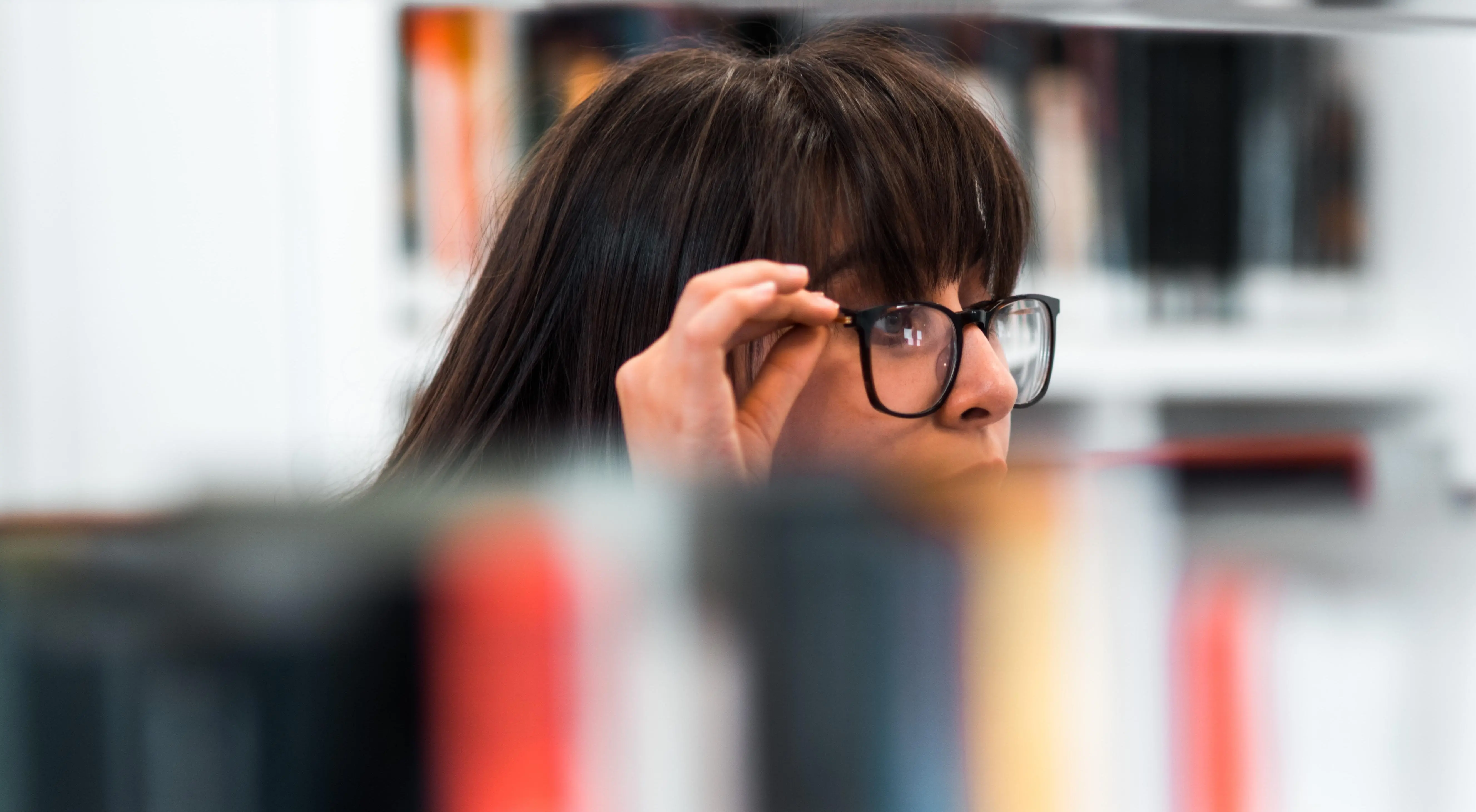 En student står bland bokhyllor och tippar upp sina svarta glasögonbågar lite